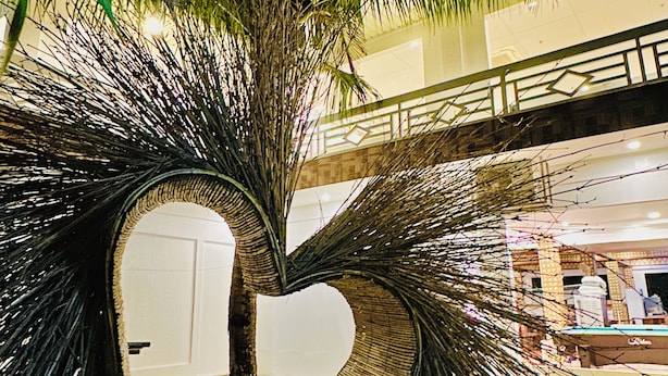 Una estructura en forma de corazón con ramas entrelazadas, rodeada de un césped cuidado y un edificio al fondo.