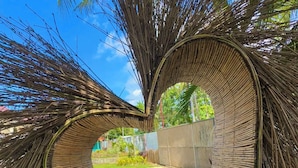 Una estructura con forma de corazón hecha de bambú, rodeada de hojas de palma, con vistas a un jardín y un edificio al fondo.