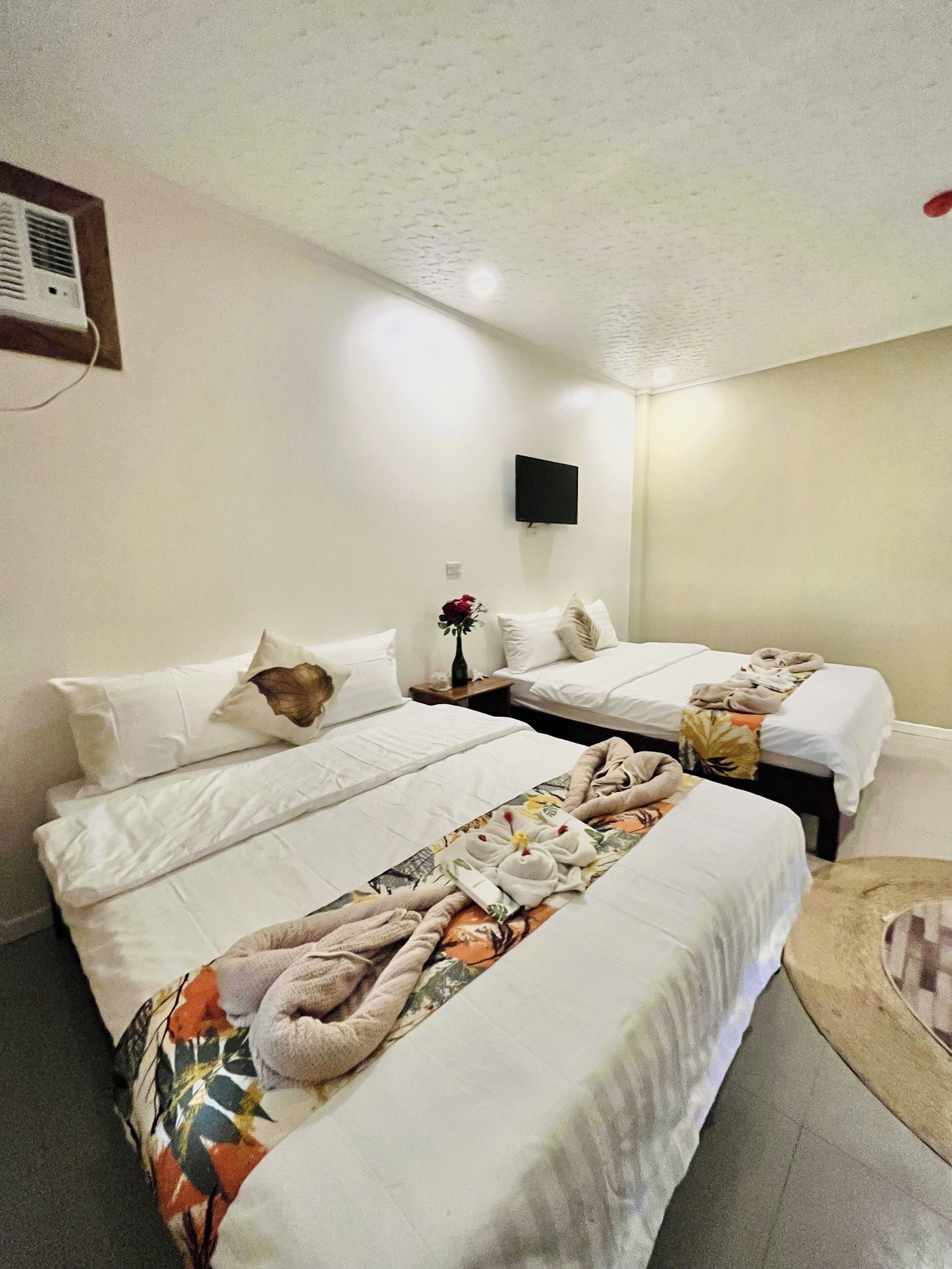 Habitación de hotel con dos camas, un televisor de pantalla plana montado en la pared y una mesita con un jarrón de flores.