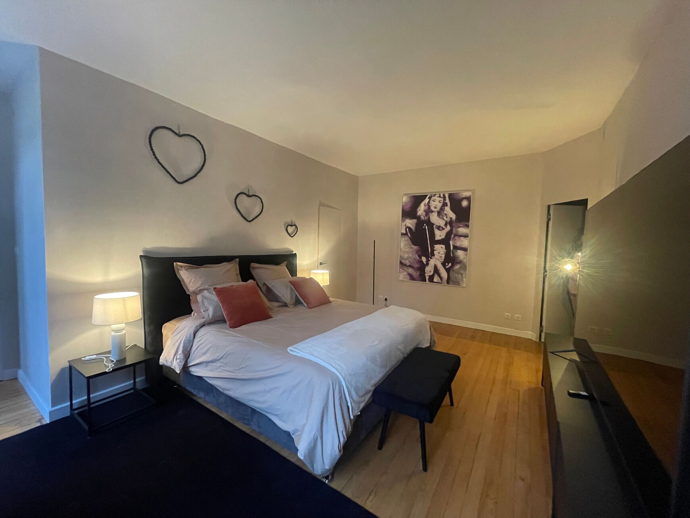 Ein modernes Schlafzimmer mit einem großen Bett, Nachttischen, einer Bank, einem an der Wand montierten Fernseher und dekorativer Wandkunst in Herzform.