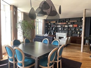 A modern dining area with a dark wooden table and blue upholstered chairs, a bookshelf filled with books and decorative items, and a fireplace in the background. - Bourgeois and modern apartment in the heart of Épinal, ideal for exploring the city (Épinal)