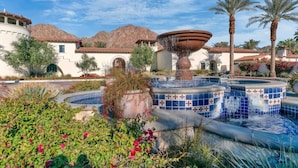 A pool area with a central fountain, surrounded by plants and palm trees, with a building in the background. - The Desert Loft B | Studio Legacy Villa (La Quinta)