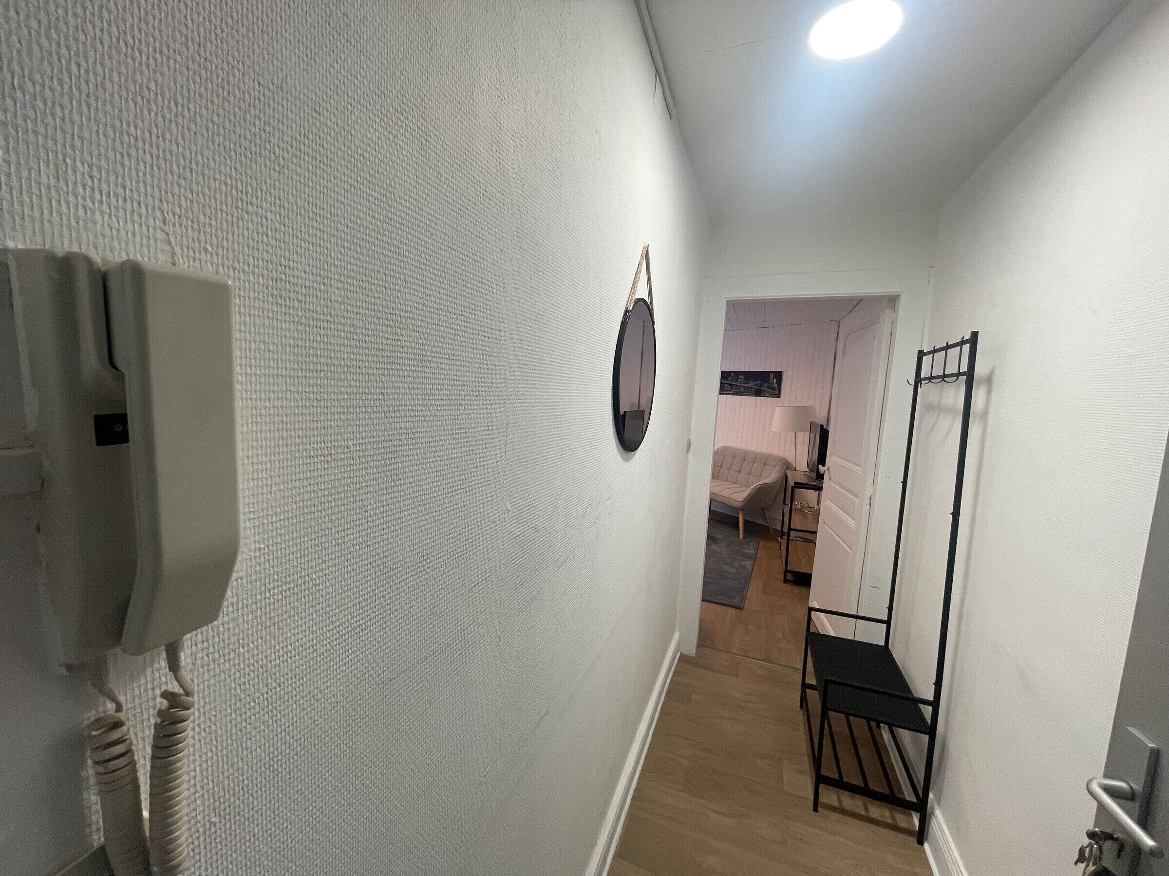 Un pasillo estrecho con paredes blancas y texturizadas, un perchero, una mesita con una silla y una puerta que da a otra habitación.
