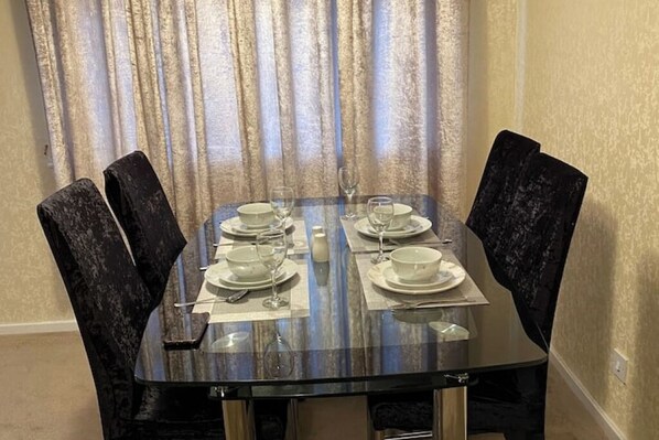 Een eettafel voor vier personen, met stoelen, een glazen blad en een gordijn op de achtergrond.