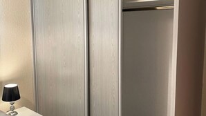 A room with a white wardrobe, a bedside table with a lamp, and a plain wall.