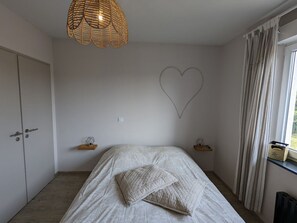 A bedroom with a bed, a hanging light fixture, a heart-shaped wall decoration, and a window with curtains.