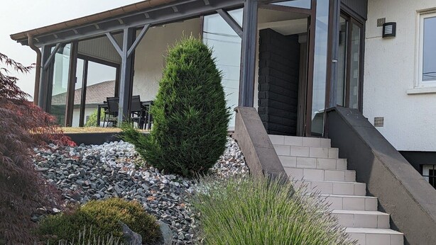 A modern house with a covered porch, a staircase leading to the entrance, and a well-maintained garden with various plants and a small tree. - Spacious house with spa, pets allowed, near Épinal (Dounoux)
