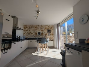 A modern kitchen with stone wall backsplash, a dining area with chairs, and a large window providing natural light. - Authentic Charm in Berry, 11 Pax, Garden and Wifi (Nohant-Vic)
