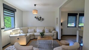 A modern living room with a sectional sofa, a coffee table, and two armchairs. There is a pendant light, a wall art piece, and a view of the outdoors through the windows. - Spacious lodge, modern comfort, peaceful garden (Saulcy-sur-Meurthe)