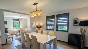 A modern living room with a dining area, featuring a large white table, beige chairs, and a hanging light fixture. - Spacious lodge, modern comfort, peaceful garden (Saulcy-sur-Meurthe)