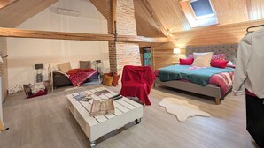 Un dormitorio tipo loft con techo inclinado, vigas de madera a la vista, pared de ladrillo y un tragaluz pequeño. Hay dos camas con distintos estilos de ropa de cama y una mesita de café blanca con libros y una bandeja.