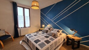 A bedroom with a large bed, a bedside table with a lamp, a window with curtains, and a geometric-patterned wall.