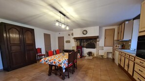 A dining area with a table covered in a patterned tablecloth, surrounded by wooden chairs. A stone fireplace is visible in the background, and a clock is mounted on the wall above it.