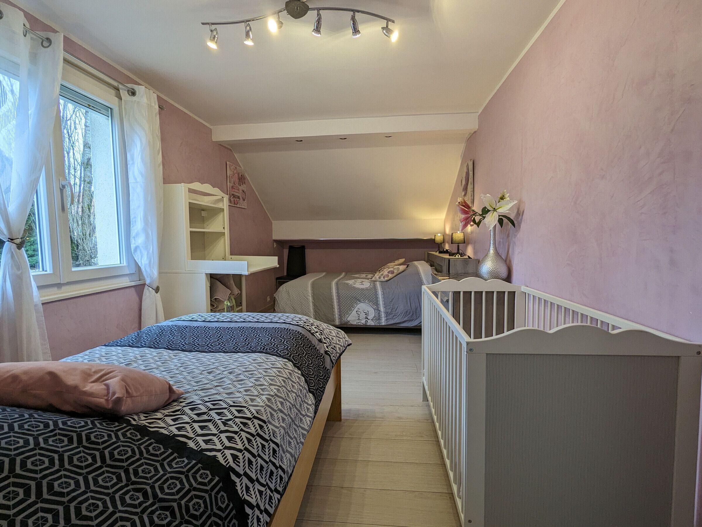 Un dormitorio con dos camas, una cuna, una ventana con cortinas, una estantería y una lámpara en el techo.