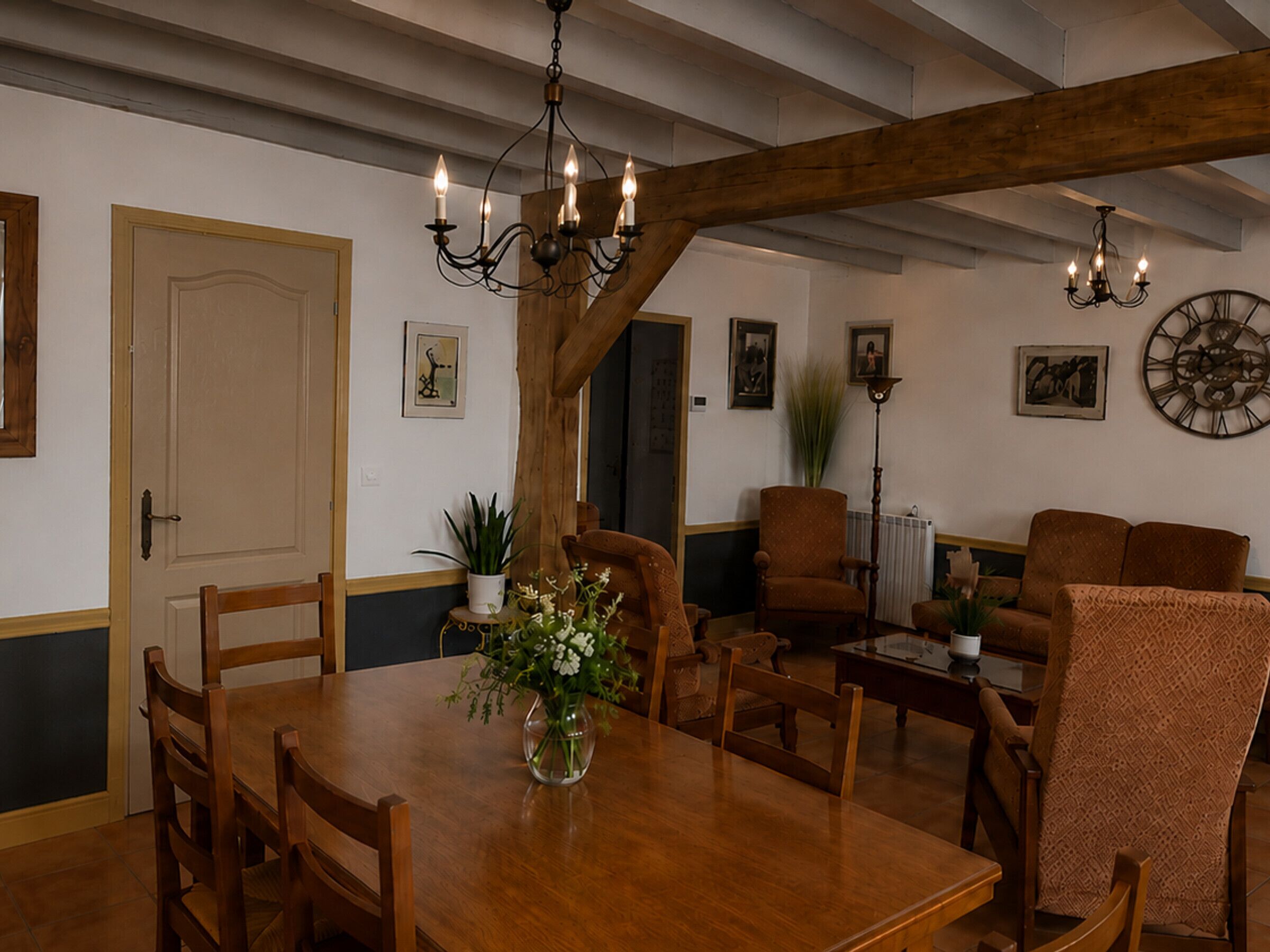 A dining area with a wooden table, chairs, a vase of flowers, a chandelier, and a clock on the wall.