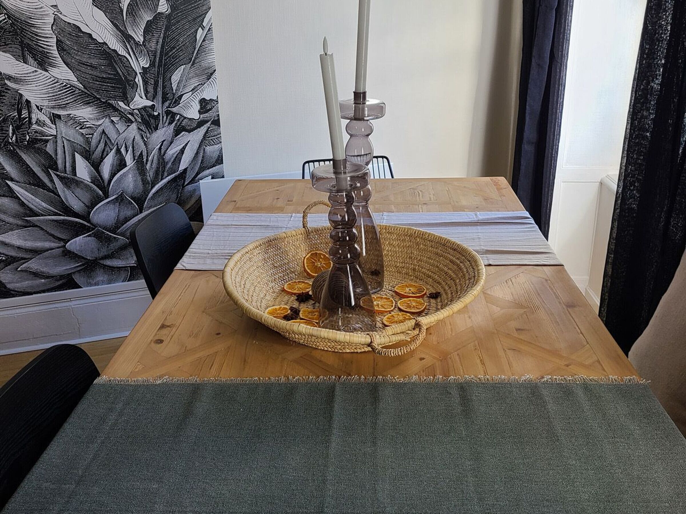Uma mesa de jantar com um arranjo de cesta trançada contendo um castiçal, fatias de laranja seca e um vaso de vidro.