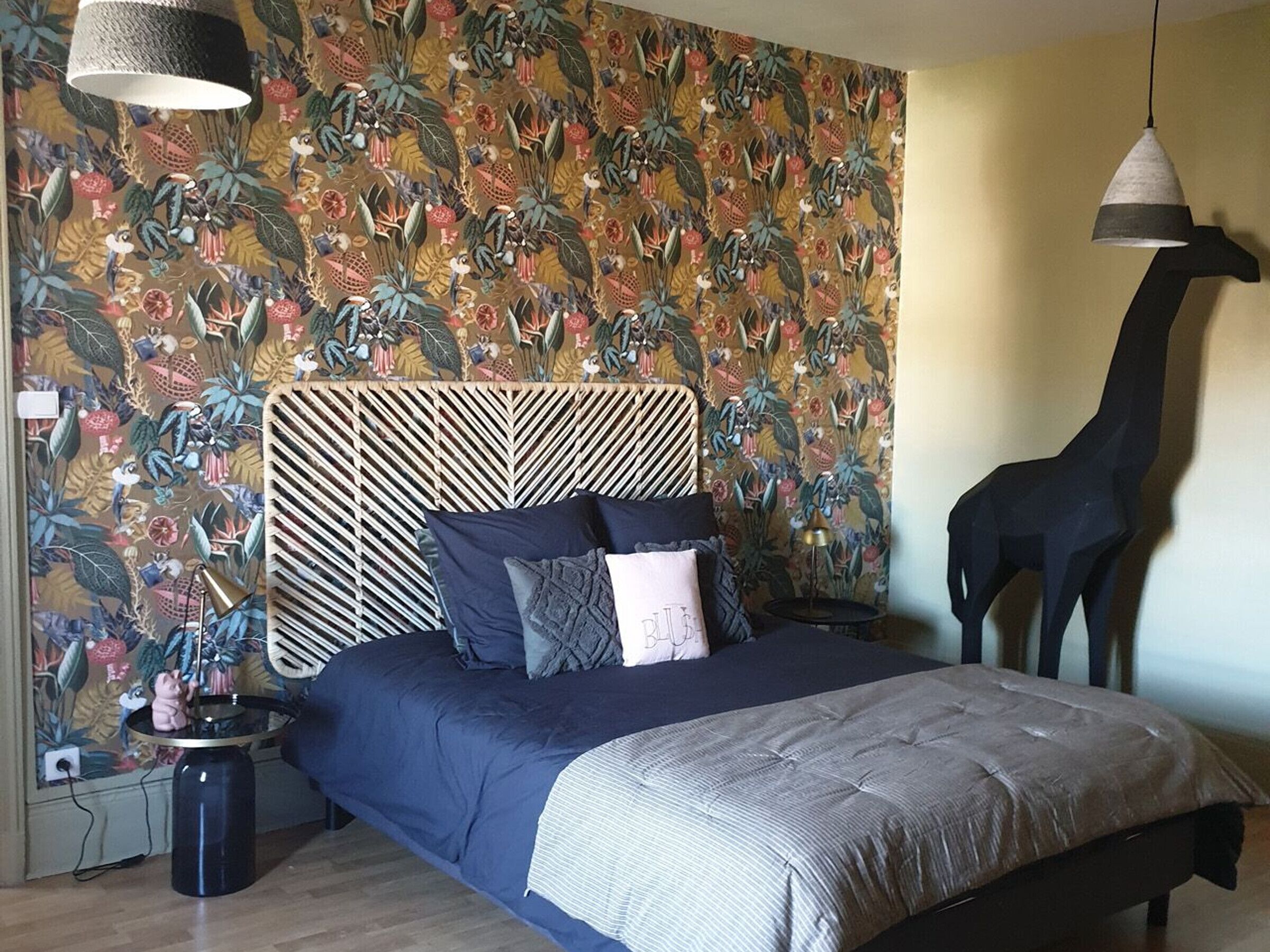 A bedroom with a patterned wallpaper, a bed with a striped headboard, a giraffe sculpture, and a hanging lamp.