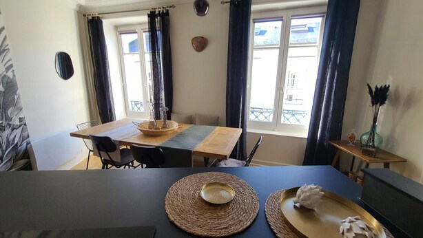 A dining area with a table set for two, a window with curtains, and a small side table with a vase.