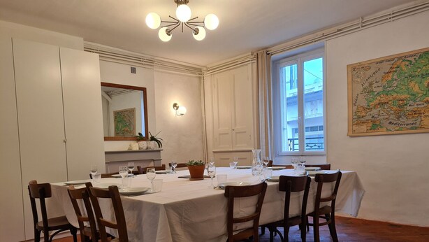 Une salle à manger avec une grande table dressée pour six personnes, entourée de chaises en bois. Il y a une fenêtre avec des rideaux, une carte accrochée au mur et un luminaire au plafond.