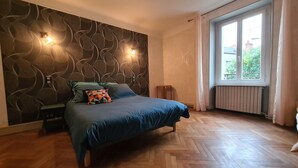 Une chambre avec un parquet en bois à motifs chevrons, un lit avec une literie bleue, un coussin géométrique aux couleurs vives et un mur orné d’un motif ondulé.