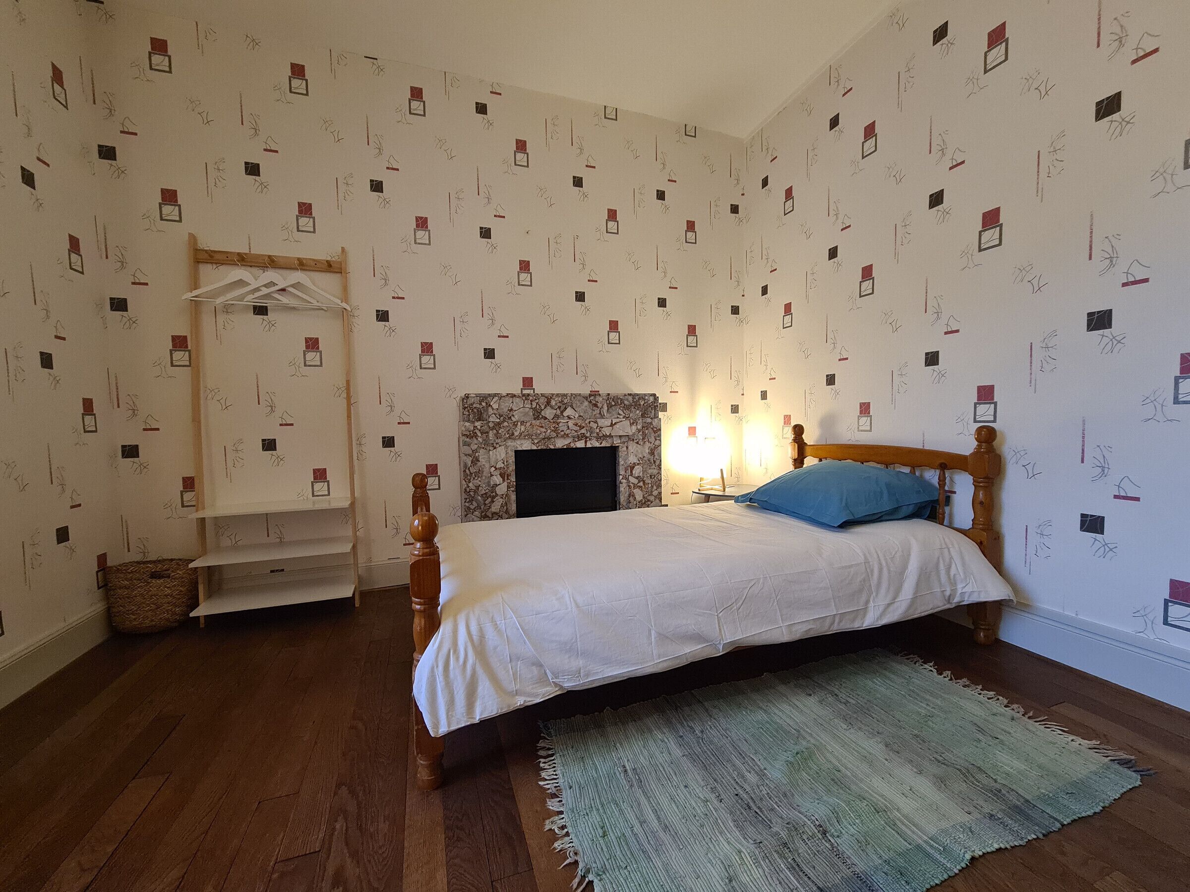 A bedroom with a bed, a wooden headboard, a fireplace, a hanging wardrobe, and a patterned wallpaper.