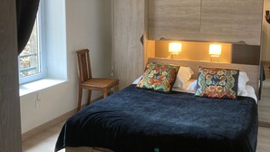 Un dormitorio con una cama, dos lámparas de noche, una silla, una ventana con cortinas y un estante en la pared con un objeto decorativo.