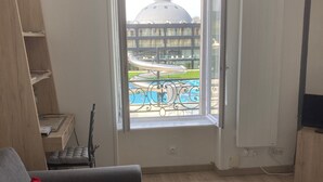 Una habitación con un sofá gris, un cojín rojo, un estante de madera y una ventana con vistas a una piscina y un edificio con cúpula.