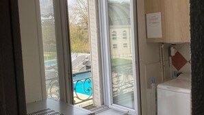 Habitación con ventana que da a una piscina y un edificio. Hay un electrodoméstico blanco en el suelo y un armario de madera encima.