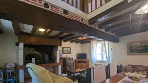 A rustic living room with exposed wooden beams, a brick fireplace, a sofa, a dining table with placemats, and a television on a wooden cabinet.