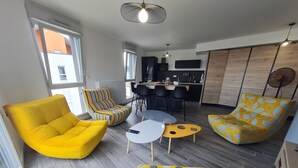 Un salon moderne avec un canapé jaune, un fauteuil à rayures et un pouf à pois. On aperçoit à l’arrière-plan un coin cuisine avec un bar et des chaises de salle à manger.