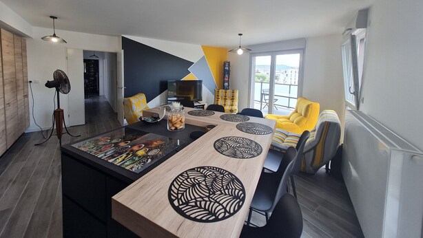 Un salon moderne avec un coin repas, comprenant une grande table au motif noir et blanc original, un canapé avec des coussins jaunes et un téléviseur fixé au mur.
