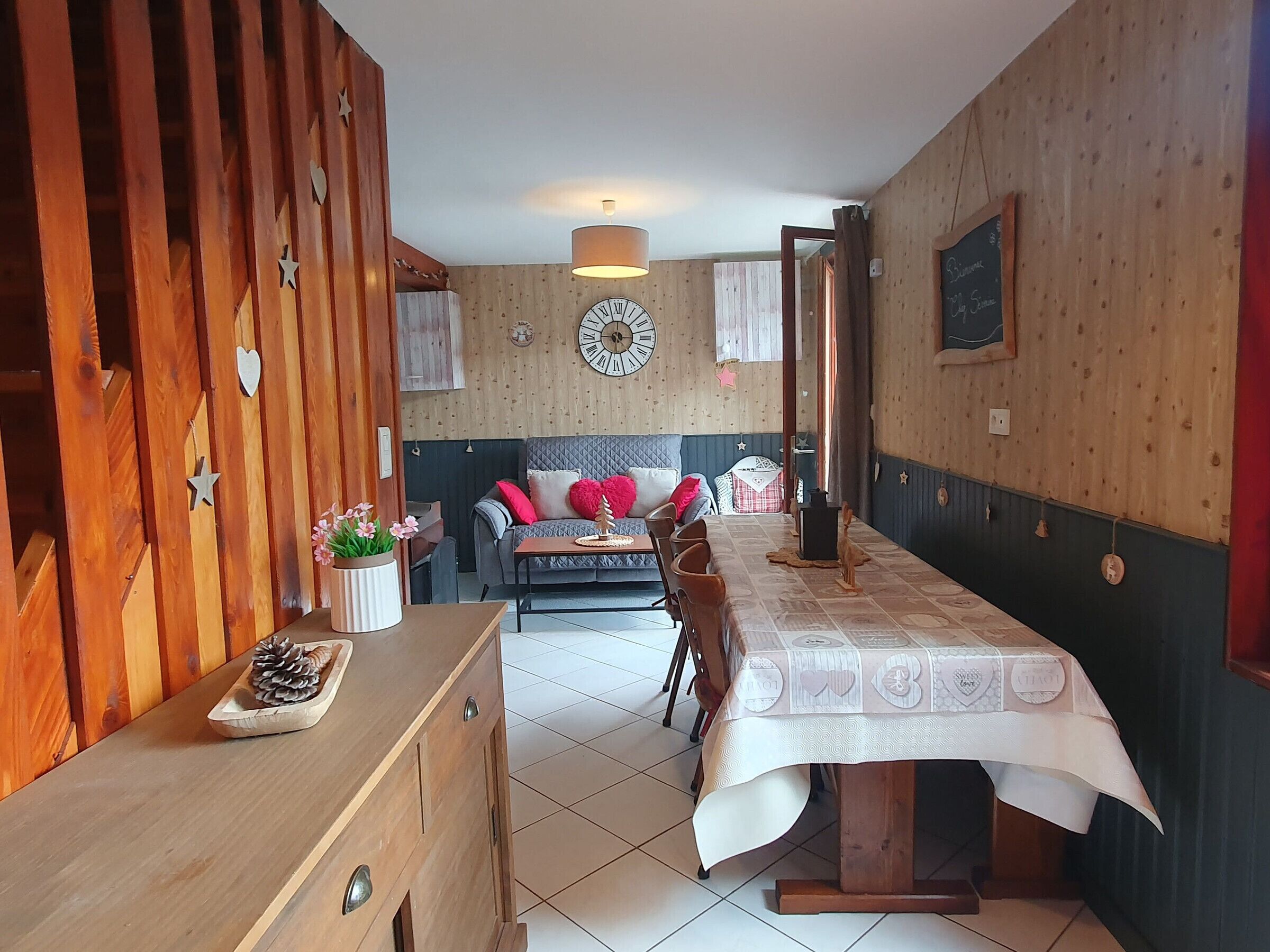 Een gezellige kamer met een houten eettafel, een bank met kussens, een wandklok en een decoratieve plant.