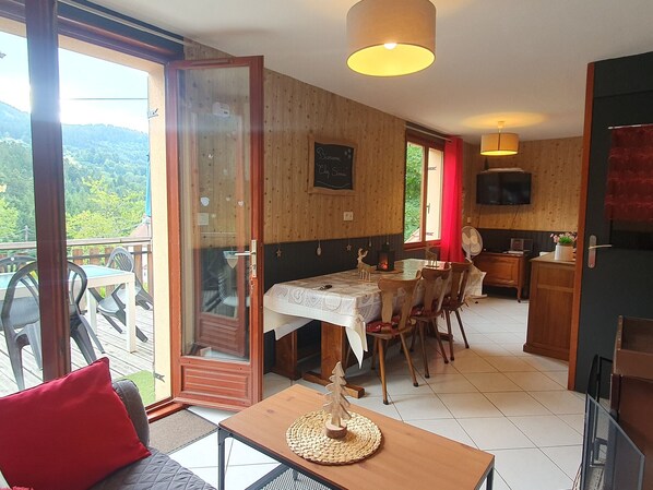 A room with a dining table, chairs, a small table with a woven placemat, a sofa with a red cushion, a television, and a wooden cabinet. - Family chalet Vosges with foosball and pets allowed (Ventron)