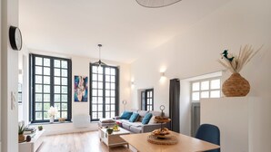 Un vaste espace de vie comprenant une grande fenêtre, un ensemble de canapés, une table à manger avec des chaises et un luminaire suspendu.