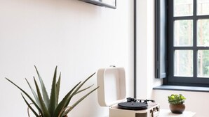 Une pièce dotée d’un téléviseur à écran plat fixé au mur, d’une fenêtre donnant sur un paysage verdoyant, d’une petite plante en pot et d’une platine vinyle posée sur une table blanche.