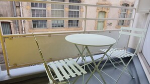 A balcony with a white table and chair, a yellow railing, and a view of a building with multiple windows and a statue. - Central and quiet apartment, near Stanislas Square (Nancy)
