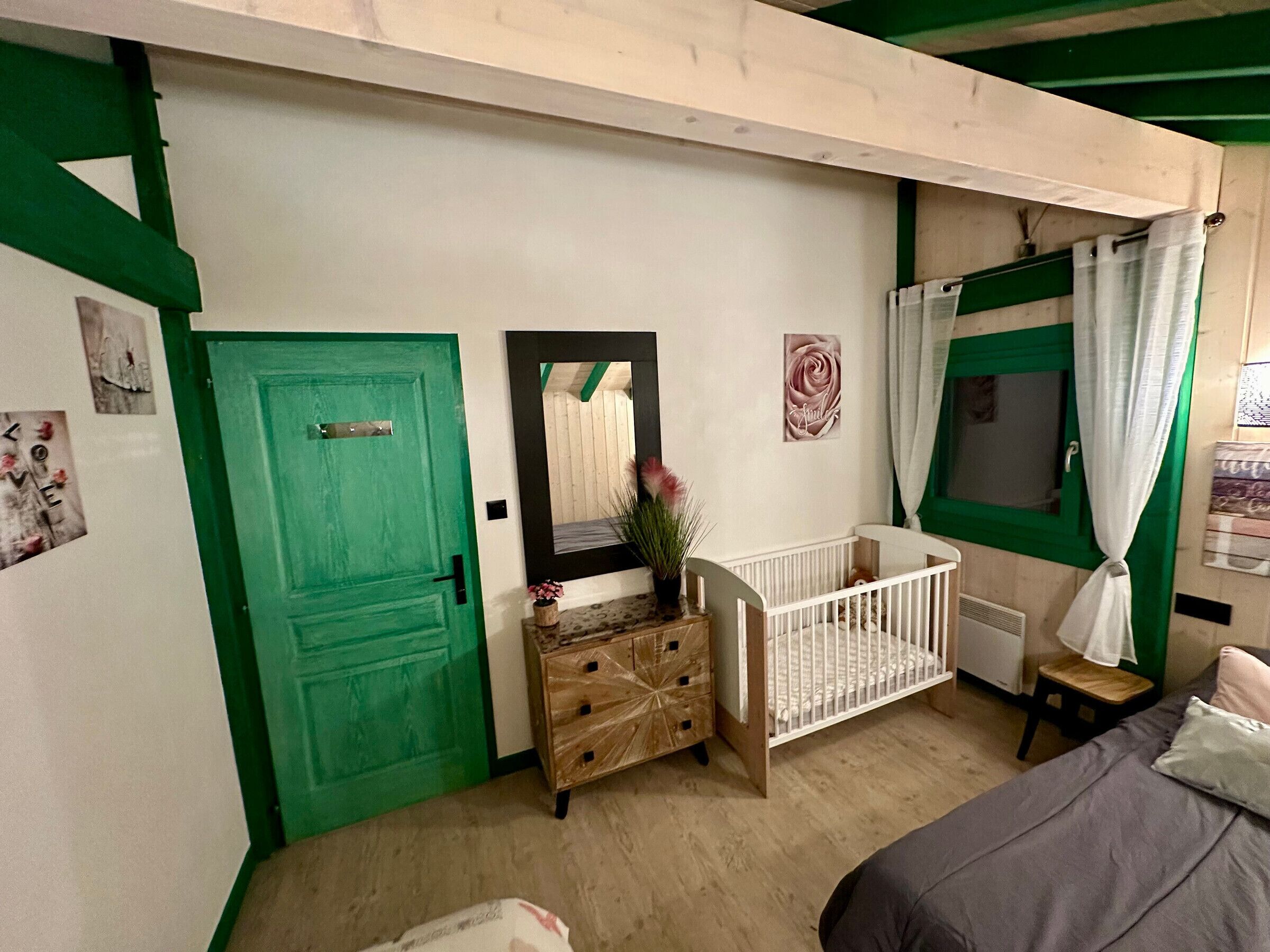 A room with a green door, a crib, a bed, and a window with curtains.