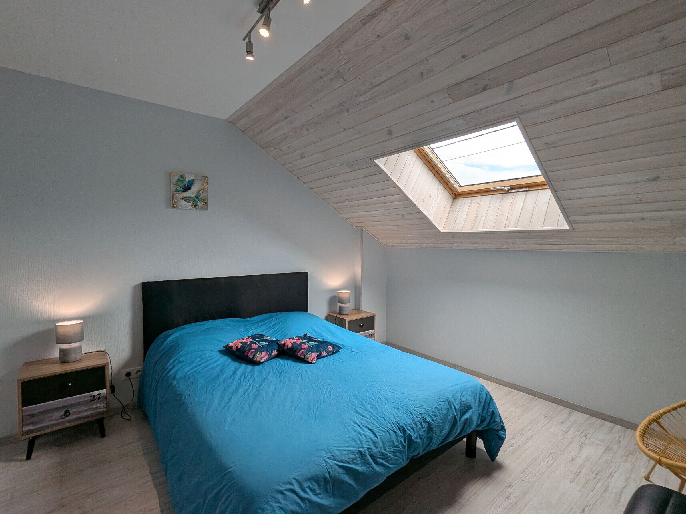A bedroom with a skylight, a bed with blue bedding, two bedside tables with lamps, and a wooden floor.