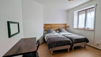 Una habitación pequeña y bien iluminada con un escritorio de madera, dos camas individuales con cabeceros de madera, una lámpara colgante y una ventana con cortinas de encaje.