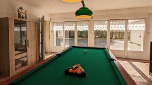 Une table de billard avec des billes, un trophée et un meuble dans une pièce dotée de grandes fenêtres et d’un luminaire au plafond.