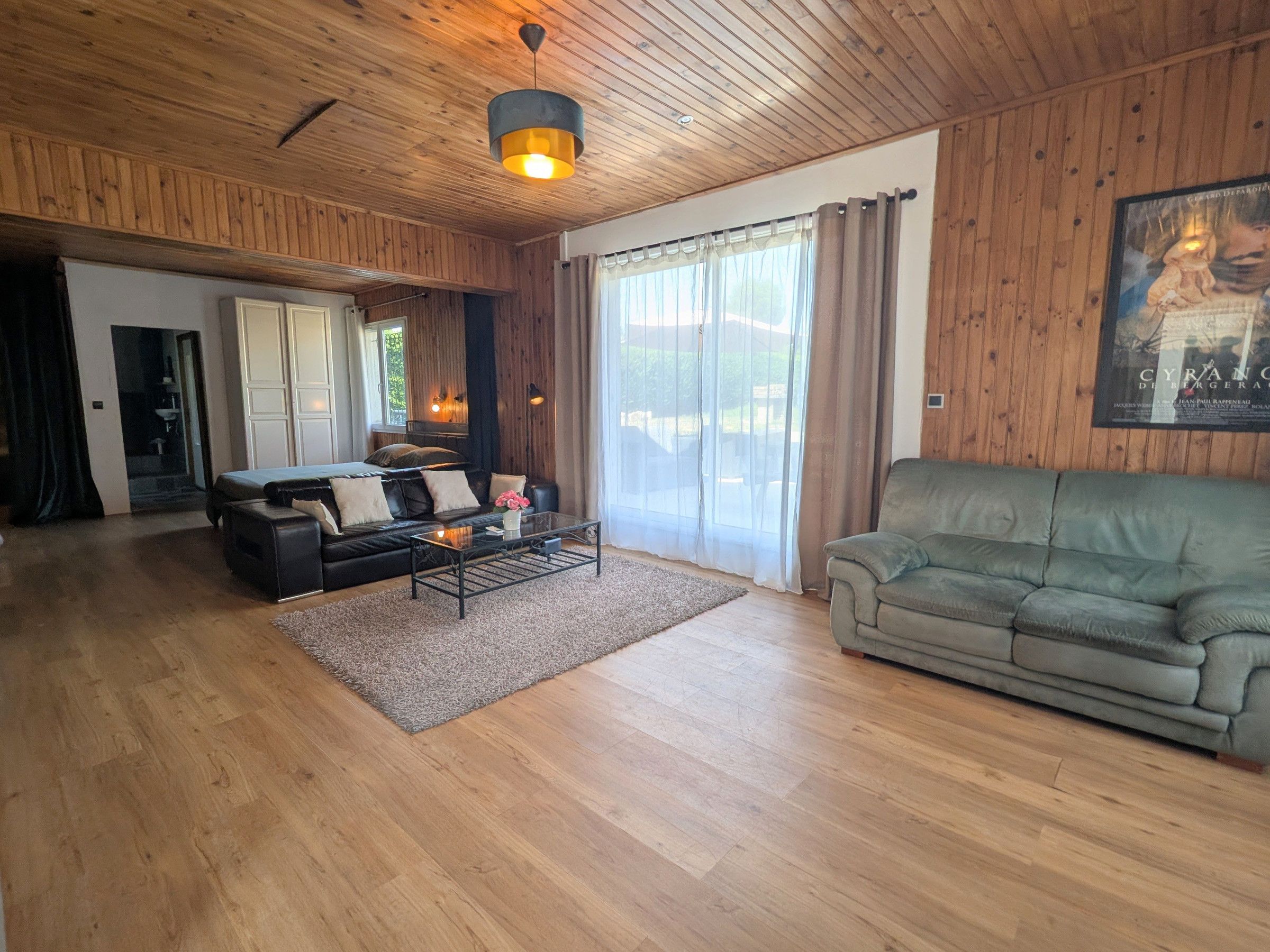 Een ruime woonkamer met een houten plafond, een bank, een salontafel en een groot raam met gordijnen.