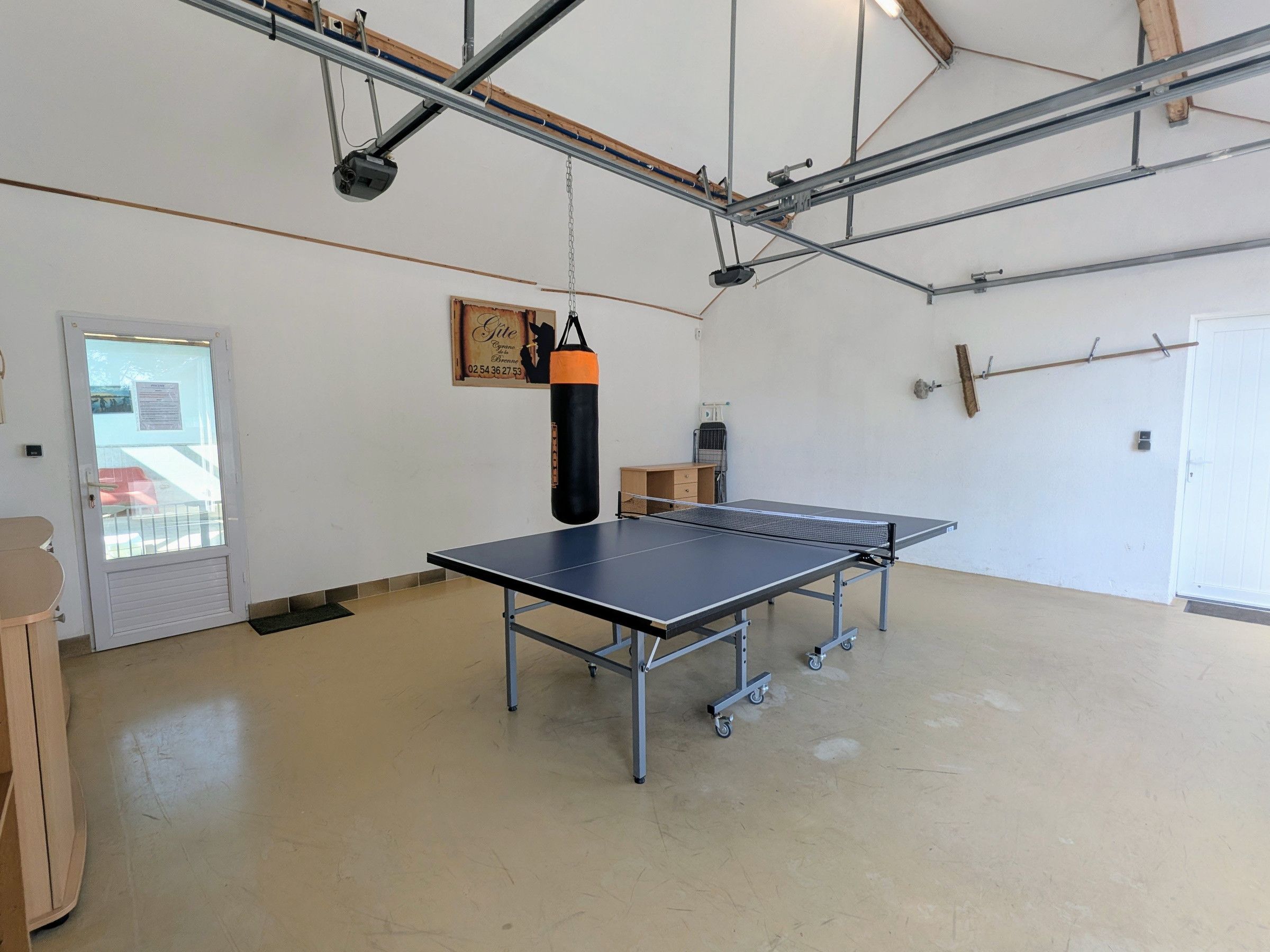 Mesa de ping pong em um quarto com uma porta, um saco de pancadas e uma estrutura metálica em forma de viga.