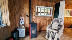 Une pièce chaleureuse aux murs en bois, dotée d’un poêle à bois, d’un fauteuil massant et d’une fenêtre avec des stores.