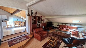 Ein geräumiger Loft mit hoher Decke, Holzbalken, einem Essbereich mit Tisch und Stühlen, einem Ledersofa, einem Fernseher und einem Bücherregal.