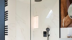 Un baño con pared de mármol, cabezal de ducha y una pequeña lámpara rectangular.