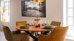Un comedor con una mesa redonda dispuesta para cuatro personas, una lámpara colgante negra y un cuadro enmarcado con una vista urbana sobre la mesa.
