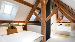 Un dormitorio con techo inclinado, vigas de madera a la vista, una cama con ropa de cama blanca, un ventanal pequeño y una puerta que da a otra habitación.