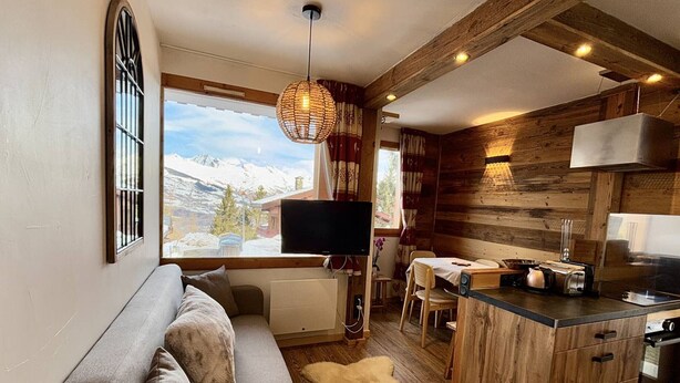 En hyggelig hytte med sofa, spiseplads, køkken og stort vindue med udsigt til et bjerglandskab.