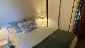En pænt redt seng med et gråt tæppe og puder, et sengebord med en lampe, og en dør til højre.