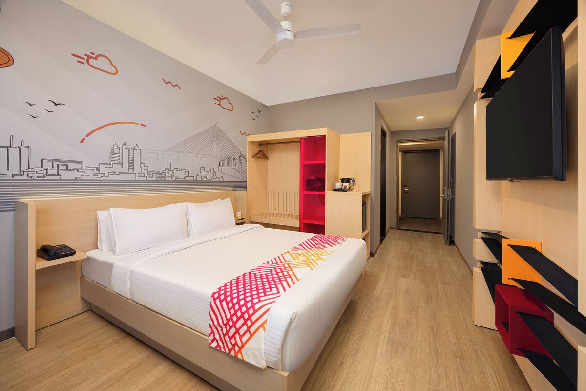 Ein Hotelzimmer mit Bett, Nachttisch, Kleiderschrank, Fernseher und Wandgemälde mit Stadtansicht.